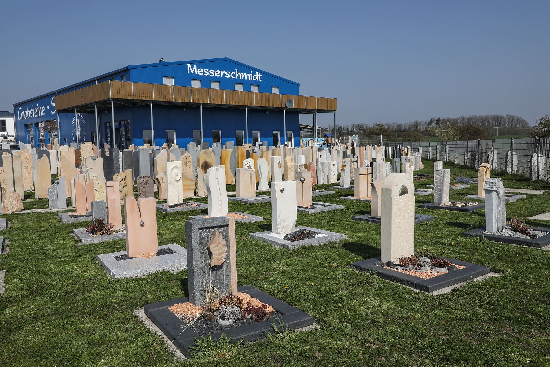 Die Ausstellung von Messerschmidt in Crailsheim: Grabsteine aus Sandstein, Kalkstein und Granit in Reihen auf der Wiese, vorne fertige Grabanlagen, dahinter die blaue Halle mit dem Schriftzug Messerschmidt