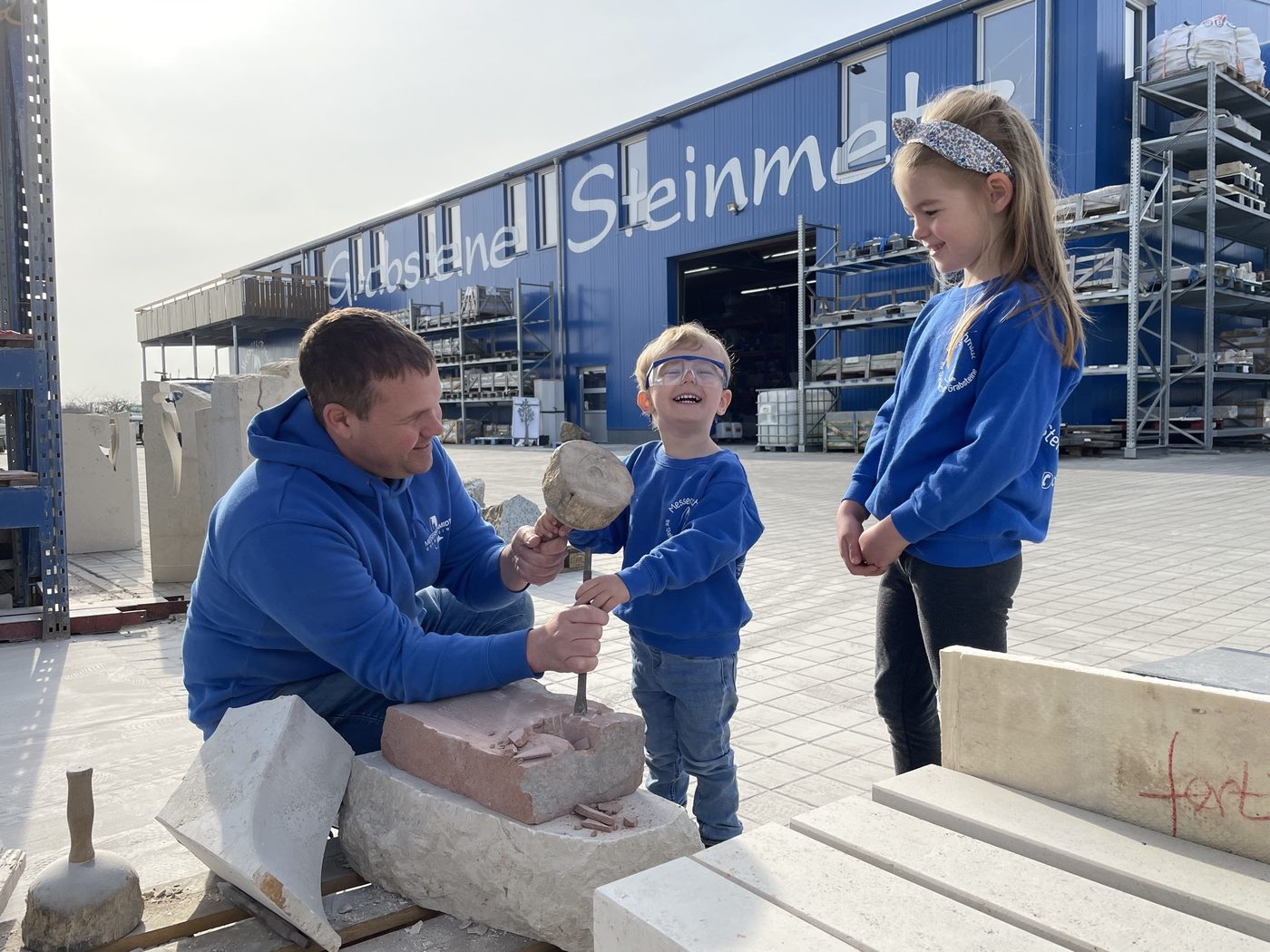 Tobias Messerschmidt arbeitet mit Hammer und Meißel an einem Stein, zwei Kinder in Firmenshirts schauen zu